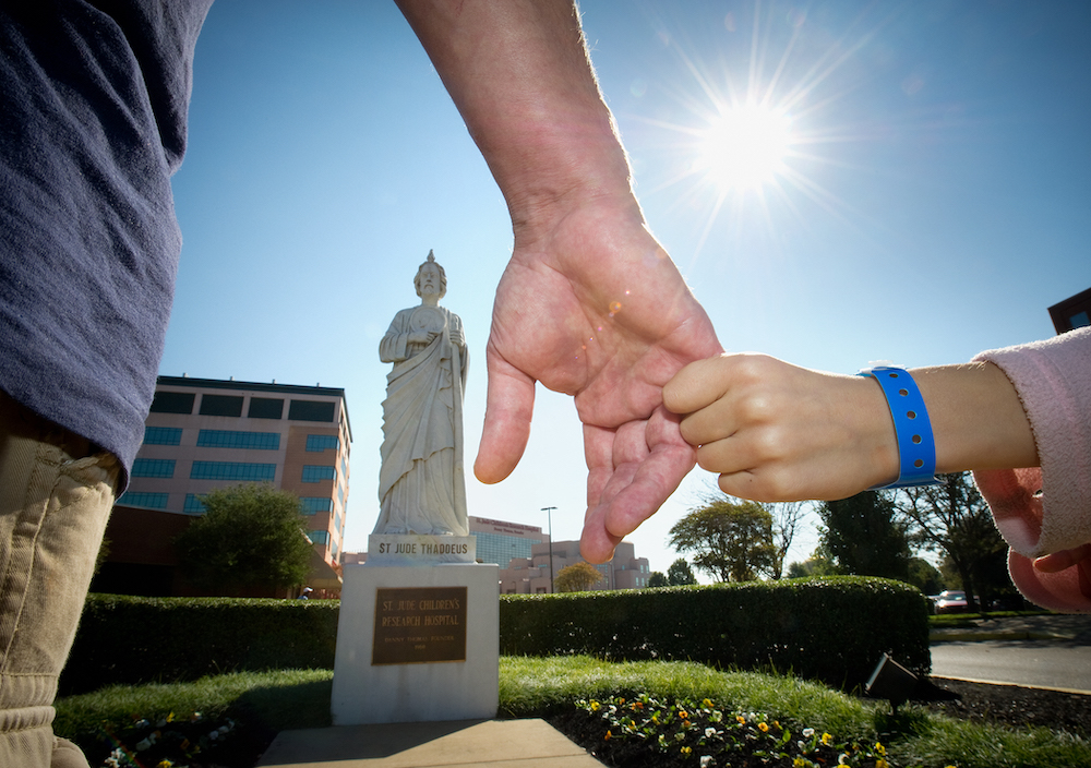 close-up of a man's hand and a child's hand (with a hospital bracelet) in front of a statue of St. Jude on the St. Jude Children's Research Hospital campus