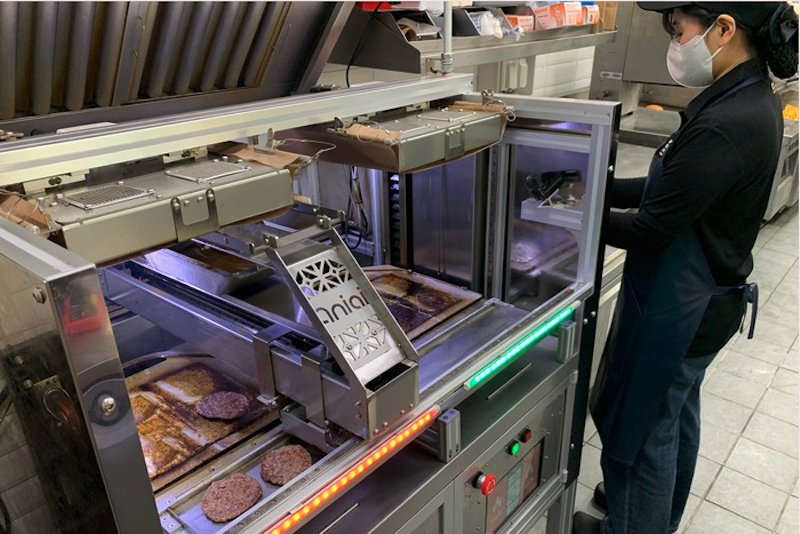 a young woman in a black uniform stands next to an automated burger-making machine