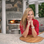 Donatella Arpaia, wearing a red chef coat, leans on a counter and smiles, with her pizza oven in the background
