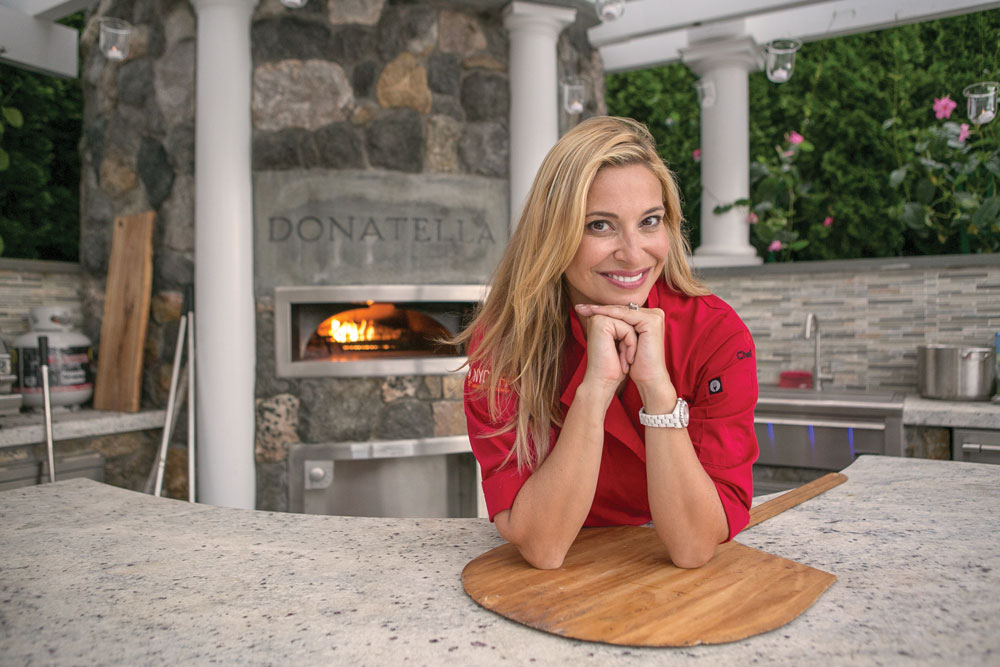 Donatella Arpaia, wearing a red chef coat, leans on a counter and smiles, with her pizza oven in the background