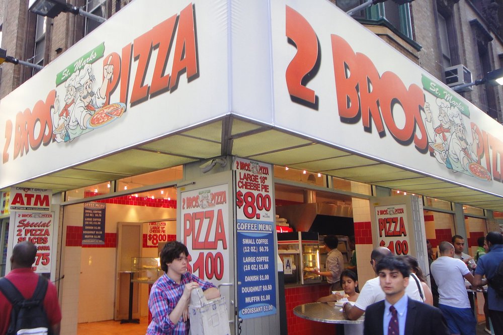 this photo shows passersby walking in front of the exterior of a 2 Bros. Pizza location in New York City.