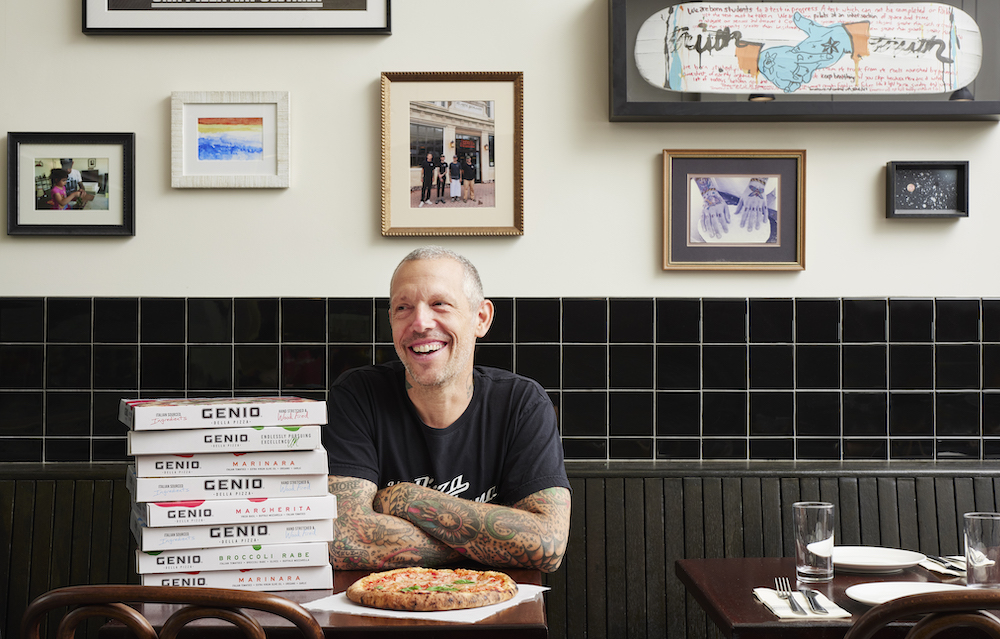 here we see Anthony Mangieri, smiling and looking away from the camera, wearing a black t-shirt and sitting at a table in his restaurant with a stack of Genio Della Pizza boxes nearby