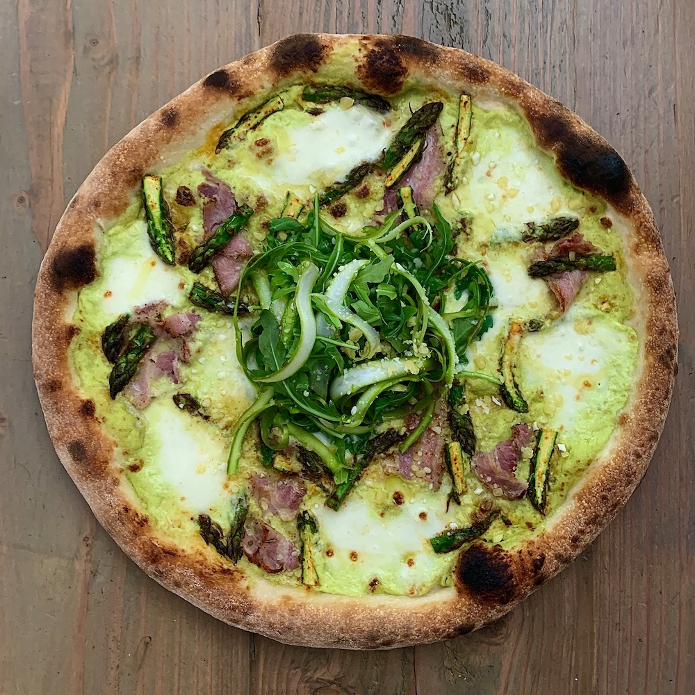 this photo shows a pizza topped with an asparagus sauce, pancetta and a big heap of wild arugula in the center