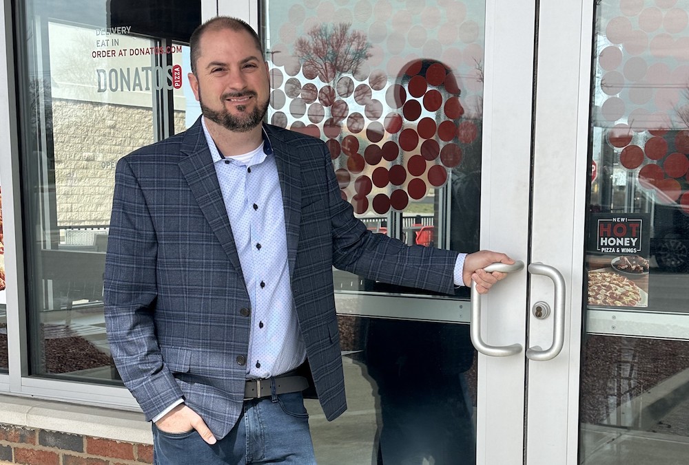 Bill Assere, wearing jeans, a light blue shirt and a blue sports coat, stands in front of a Donatos restaurant with his hand on the front-door entrance