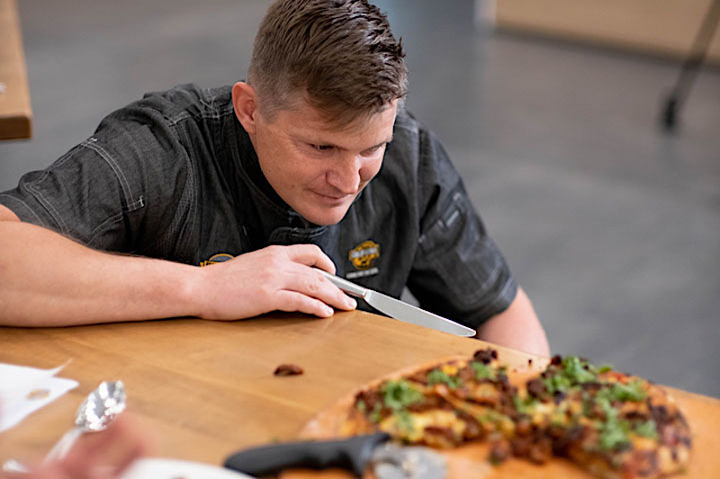 this photo shows a pizza chef in a dark chef coat kneeling down and peering closely at a pizza