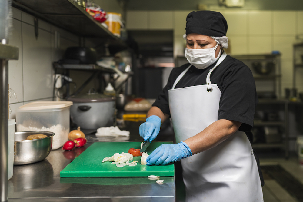 this shows a woman wearing a COVID mask, a black shirt and white apron cutting onions on a plastic board in a restaurant kitchen