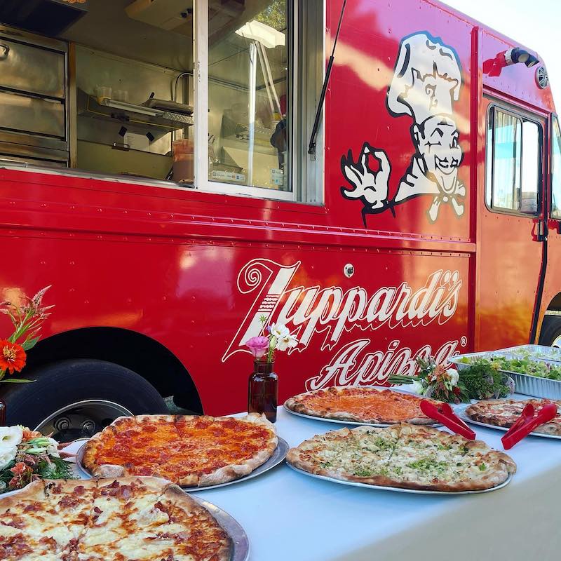 this photo shows an array of pizzas on a table next to the logoed Zuppardi's food truck