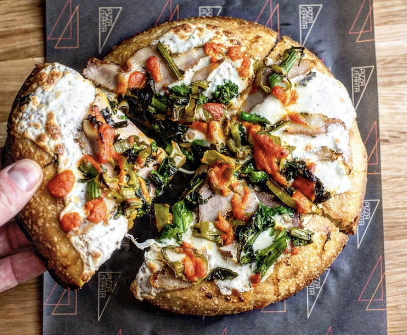 Here'a beautiful pizza featuring porchetta, mozzarella, broccoli rabe and other ingredients. A hand is pulling a slice from the whole pie, which is personal-sized.