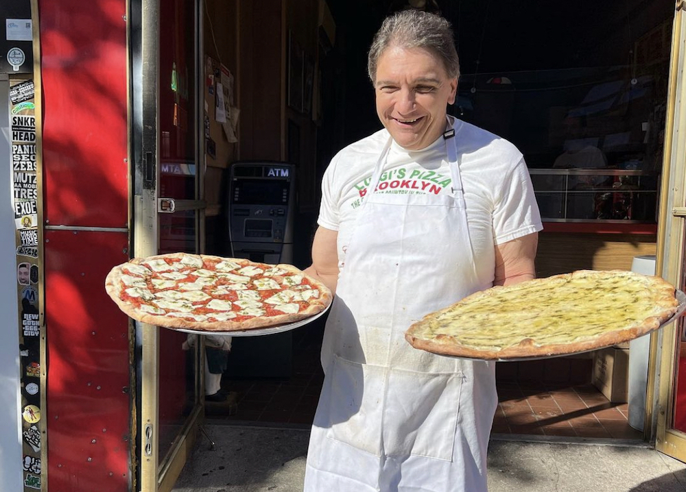 this shows Giovanni Lanzo holding two New York-style pizzas in front of Luigi's Pizza. He's wearing a white Luigi's-logoed T-shirt, a white apron and white shorts and has thinning, gray hair