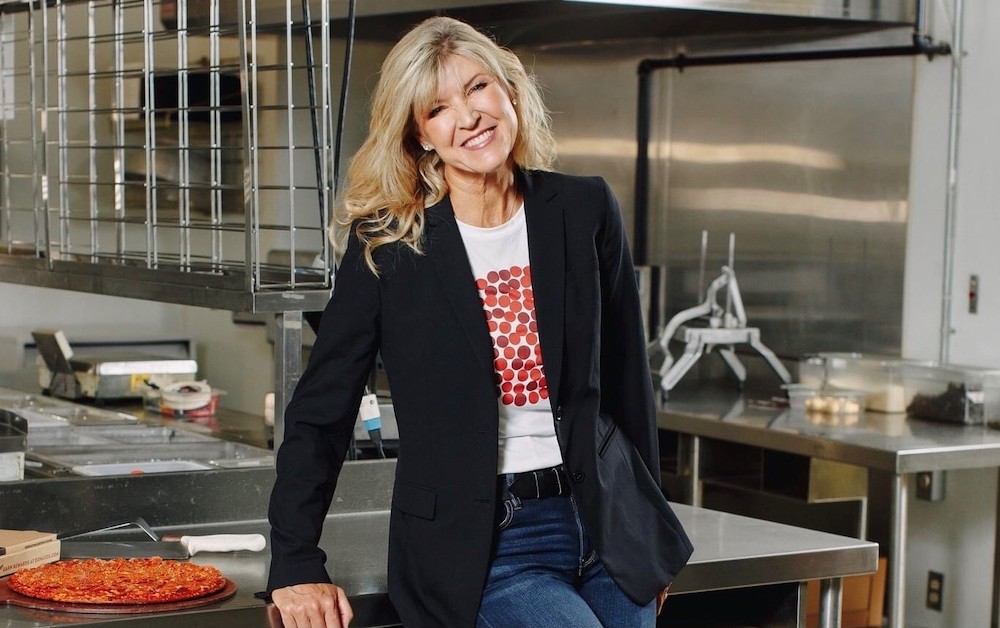 this photo shows Jane, with long blonde hair, a dark jacket over a white t-shirt and blue jeans, leaning against a prep table with a pepperoni pizza next to her on the table