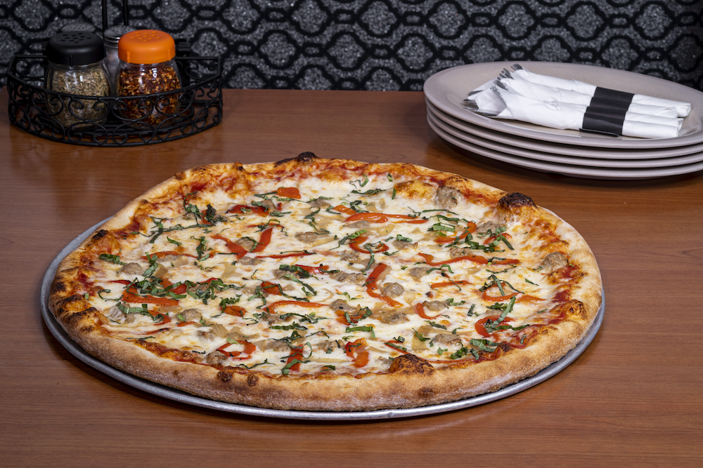 a whole pizza topped with crumbled sausage, smoked mozzarella and a sriracha-infused sauce, set on a brown table with several stacked plates in the background