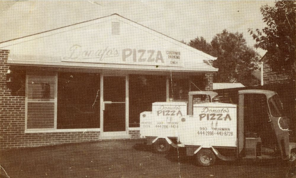 this is a vintage black-and-white photo of the original Donatos location, a brick building with a pair of compact trucks in front bearing the Donatos logo, address and phone number