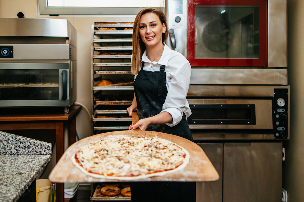 dressed in a white uniform shirt and dark apron, a woman in a pizza kitchen displays a pizza before placing it into the oven