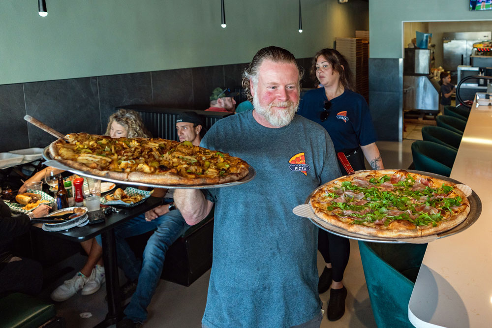 this photo shows a man with a grey beard and a blue shirt carrying two large pizzas through Madison Avenue Pizza