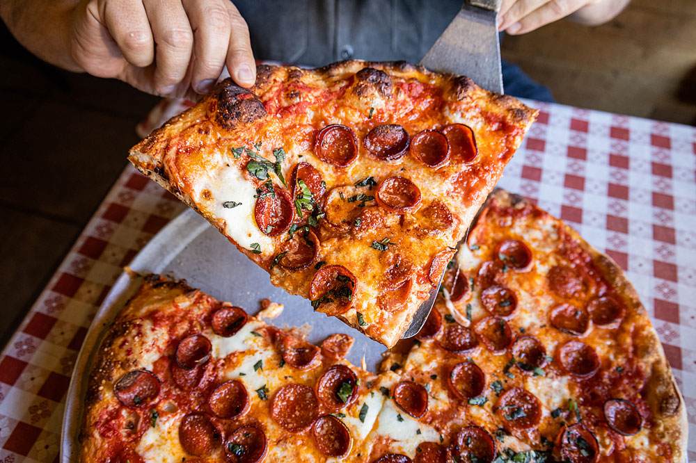 this shows a person's hand holding up a slice of pizza with pepperoni, mozzarella and shreds of basil