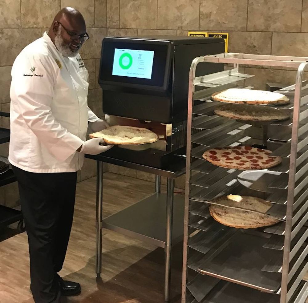 this photo shows an African-American chef in a white chef coat holding a cheese-topped pizza next to the xPizzaCube from xRobotics