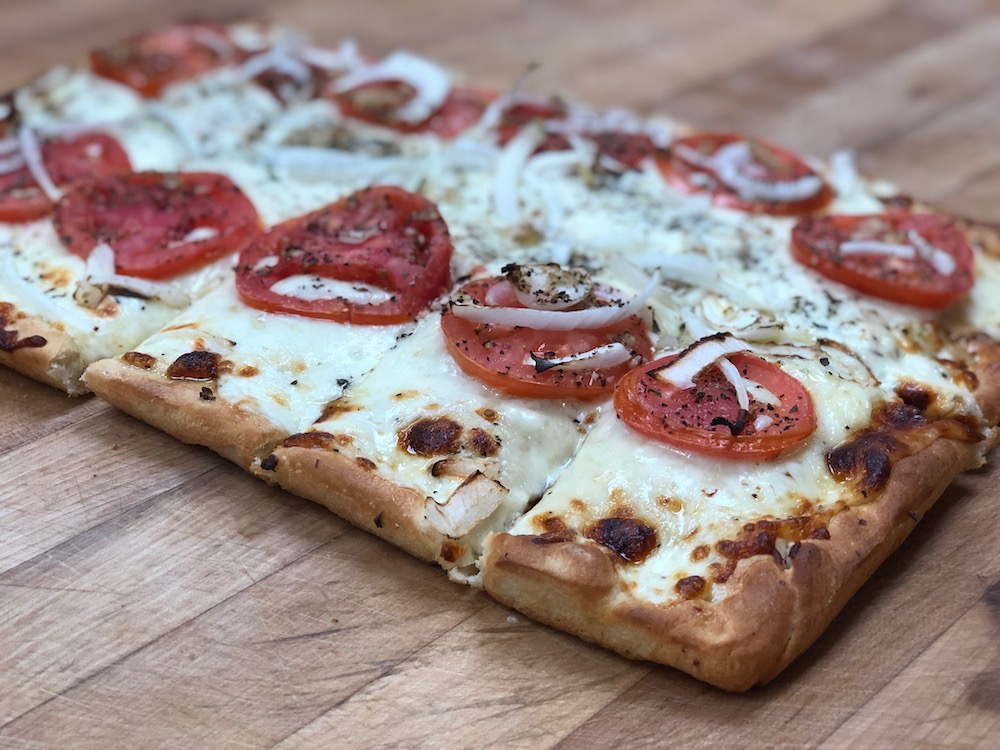 this photo shows a whole Old Forge-style pizza with a thick layer of cheese on top, tomato slices, onions and seasoning.