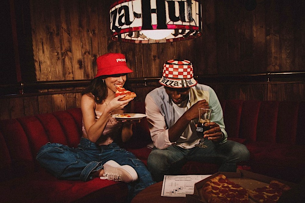this photo shows a young couple eating pizza in their home underneath a Pizza Hut lamp. One is wearing the side of the hat that resembles the Pizza Hut roof, and the other wears the side of the hat that resembles Pizza Hut's lamp shades.