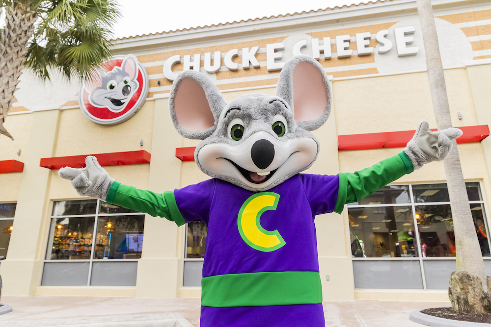 this photo shows the animatronic Chuck E. Cheese with widespread arms in a welcoming gesture in front of a Chuck E. Cheese restaurant