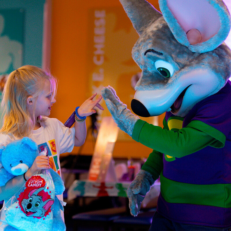 this shows an adorable little girl in a t-shirt with blonde hair high-fiving the animatronic Chuck E. Cheese