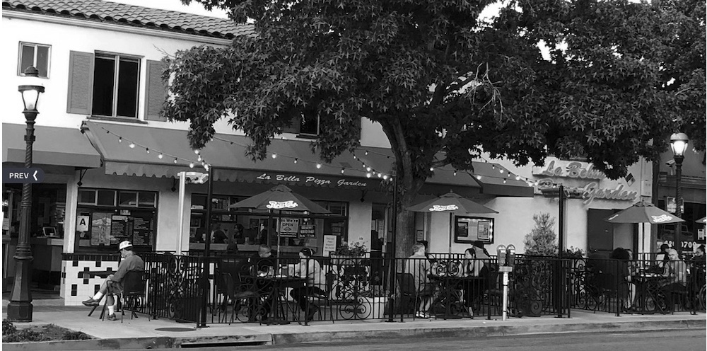 this black and white photo shows the exterior of La Bella Pizza Garden with customers sitting outside on the patio beneath a large tree