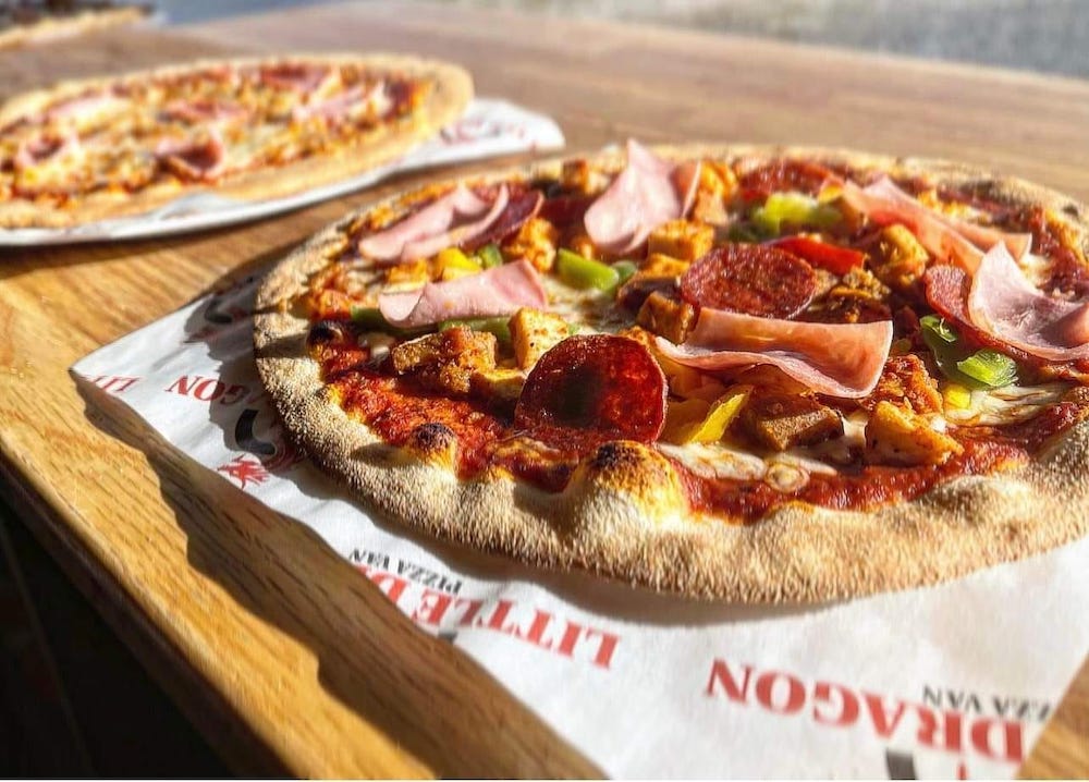 in the foreground of this photo, there's a pizza with pepperoni, ham and other meats and veggies, with a second pizza blurred in the background, both on paper logged Little Dragon Pizza Van