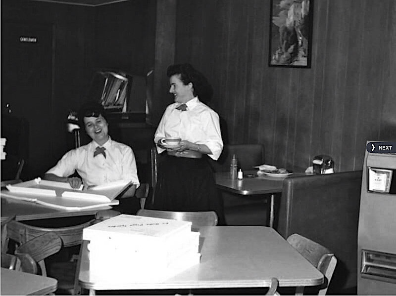 this vintage black and white photo shows two women servers in white shirts, laughing together. One is folding a pizza box and the other holds a cup of coffee