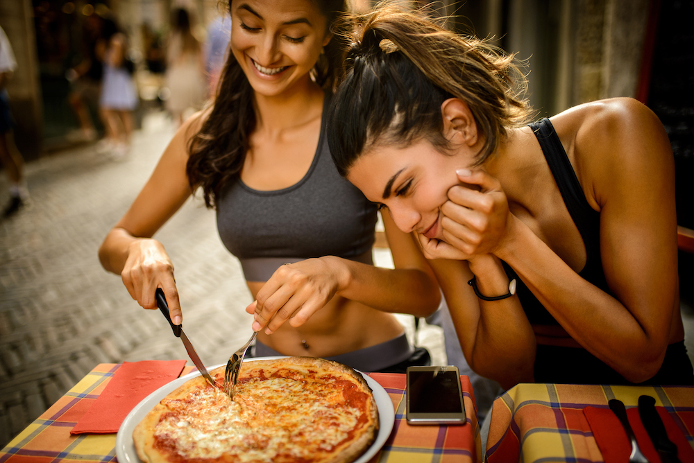 Sporty woman eating pizza while her friend is gazing at it adoringly