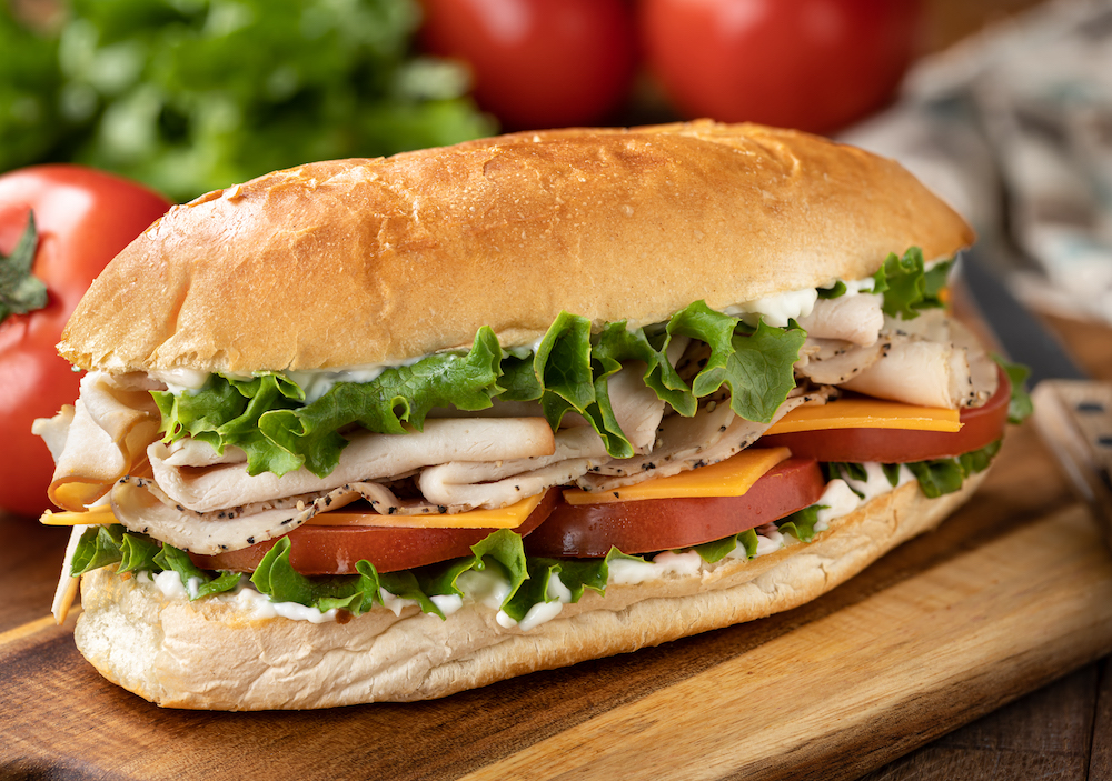 Submarine sandwich made with turkey, ham, cheese lettuce and tomato on a hoagie roll with lettuce and tomatoes in background