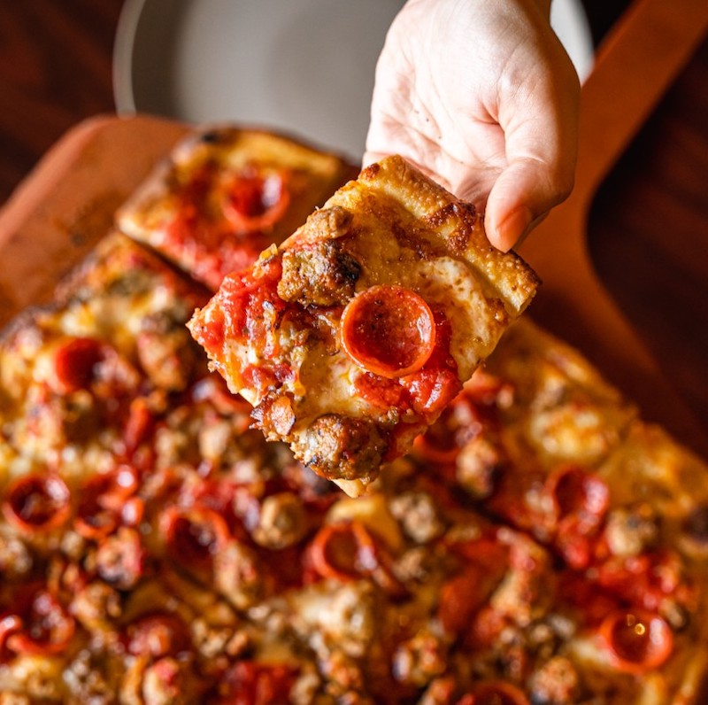 this photo shows a person's hand holding up a slice of delicious-looking pizza topped with pepperoni and sausage