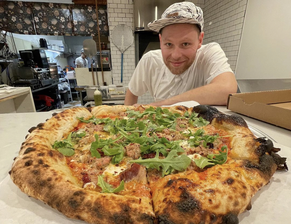 this photo shows Ben Osher grinning as he sits at a table and peers out above a large pizza with a very charred crust