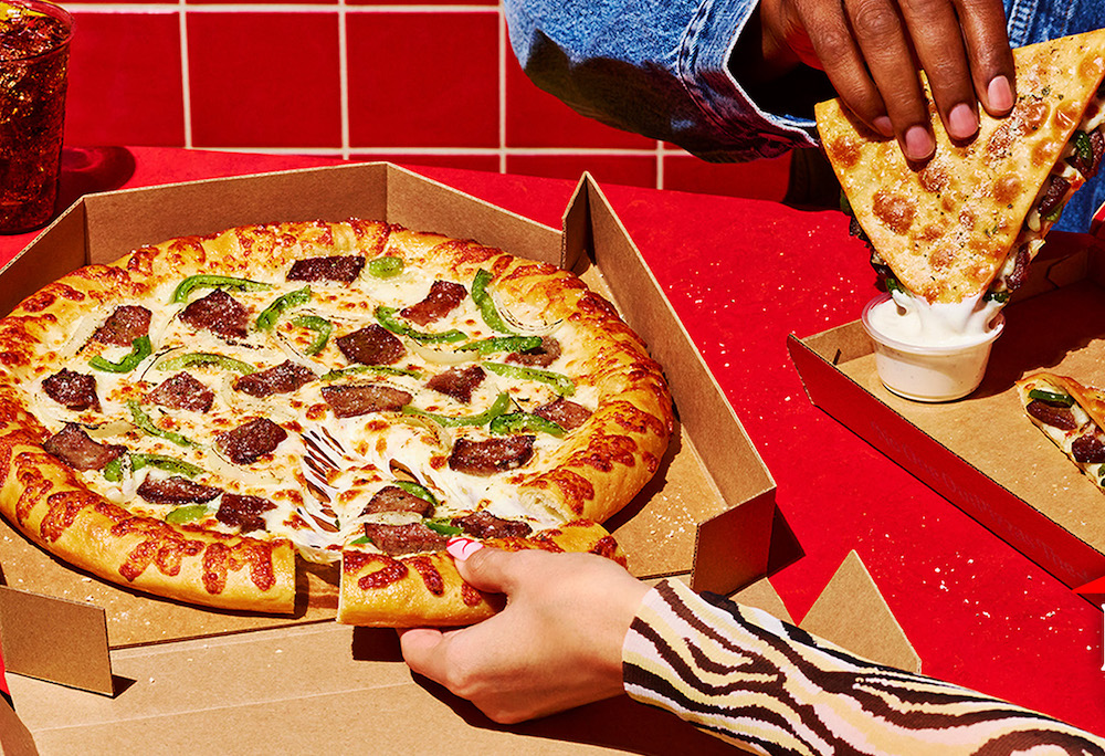 this photo shows one person's hand pulling out a slice of the cheesesteak pizza and another person's hand dipping a cheesesteak melt in ranch sauce