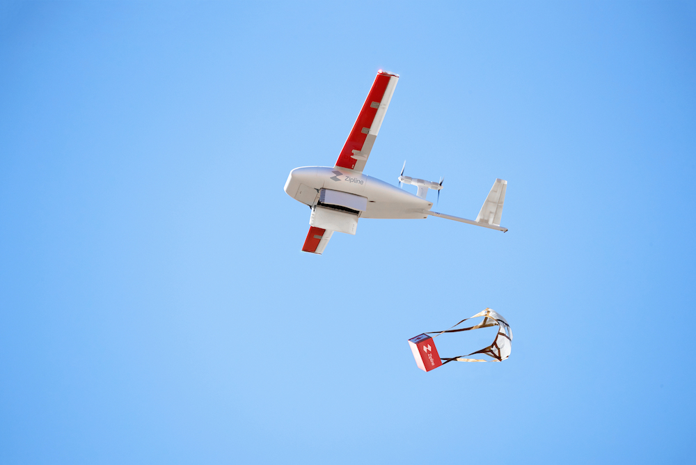 this photo shows an airborne drone, set against a bright blue sky, dropping a second smaller delivery drone to the ground.