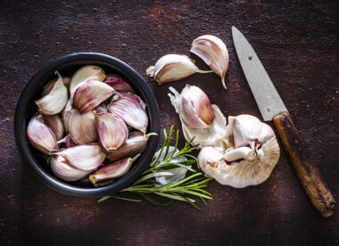 Garlic cloves in a black bowl shot from above on rustic brown background. A garlic bulb an three cloves are beside the bowl. An old kitchen knife is at the right of an horizontal frame.