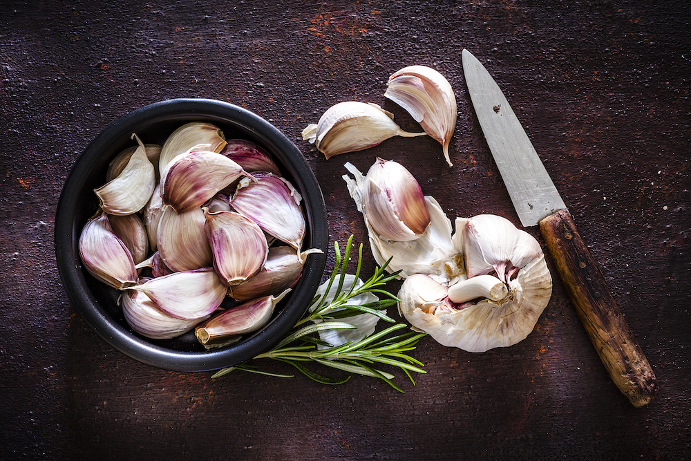 Garlic cloves in a black bowl shot from above on rustic brown background. A garlic bulb an three cloves are beside the bowl. An old kitchen knife is at the right of an horizontal frame.