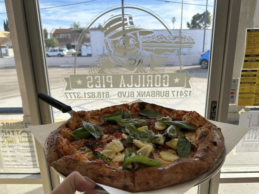 this photo shows one of Gorilla Pies' pizzas, topped with basil leaves and some kind of green topping that looks like fruit, placed on a pan in front of a window bearing the Gorilla Pies logo