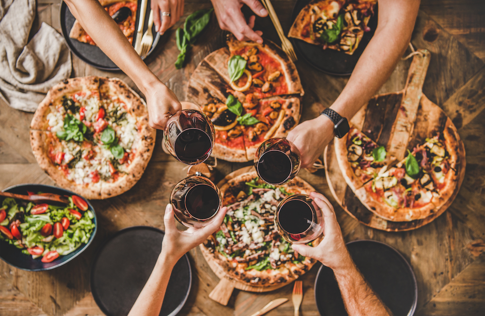 This shows an overhead view of a group of people clinking glasses with red wine over a rustic wooden table with various kinds of Italian pizza.