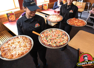 this photo shows two smiling servers bringing pizzas to customers' tables at Happy Joe's.