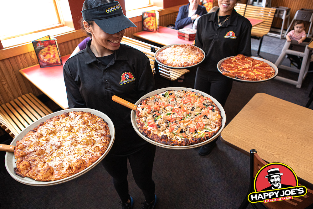 this photo shows two smiling servers bringing pizzas to customers' tables at Happy Joe's.