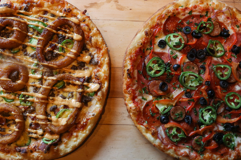 this photo shows two different pizzas, including one (on the left) topped with onion rings, jalapeños and ground sausage and the one on the right topped with pepperoni, jalapeños, black olives and sausage slices