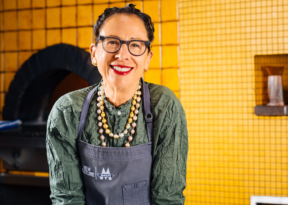 this photo shows a bespectacled Nancy Silverton in a green shirt and blue apron, smiling and leaning forward, against a yellowish background