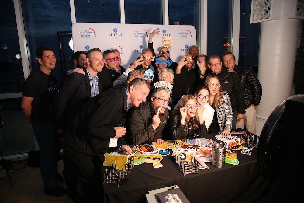 this photo shows a large group of women and men, most wearing dark clothes, wearing novelty glasses and other novelty items and gathered around a table piled with such items.