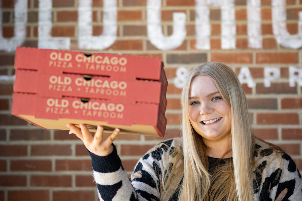 this photo shows a woman with long, straight blond hair smiling as she holds up three Old Chicago pizza boxes