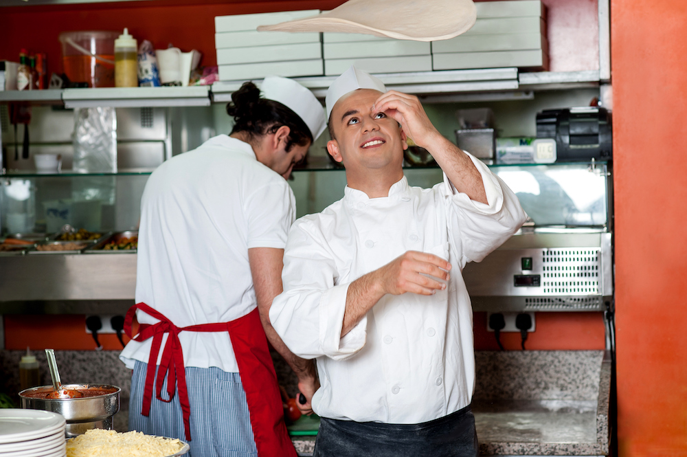 a smiling chef spins pizza dough in the air. He's wearing a white chef's coat. Behind him, another man in a white shirt can be seen working with his back towards the camera.