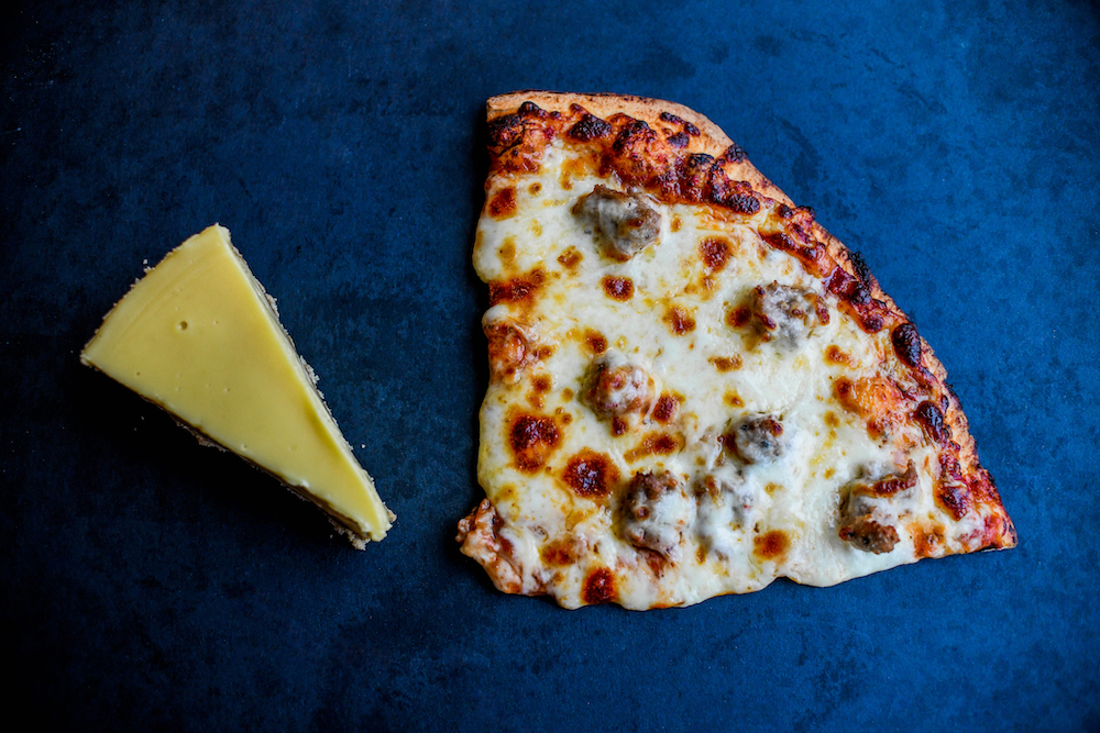 this photo shows a single slice of key lime pie placed alongside a slice of pizza against a dark blue background