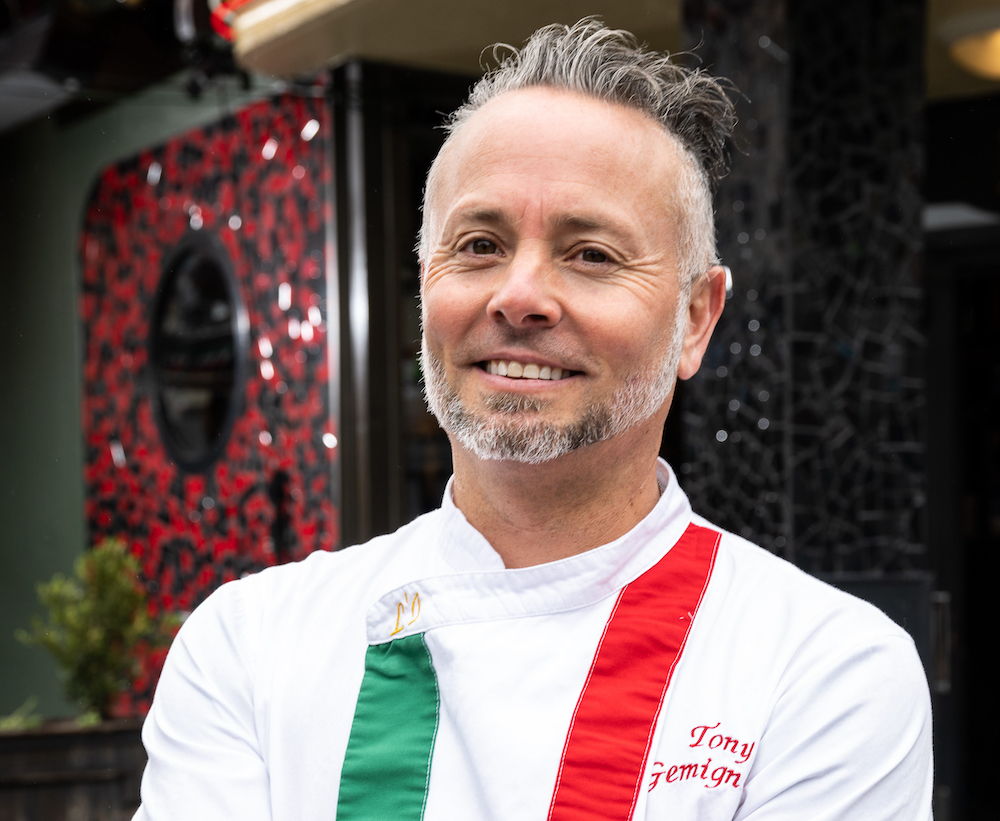 This photo is a headshot of Tony Gemignani with thinning, graying hair wearing a white chef coat with one thick green stripe on one side and one thick red stripe on the other side.