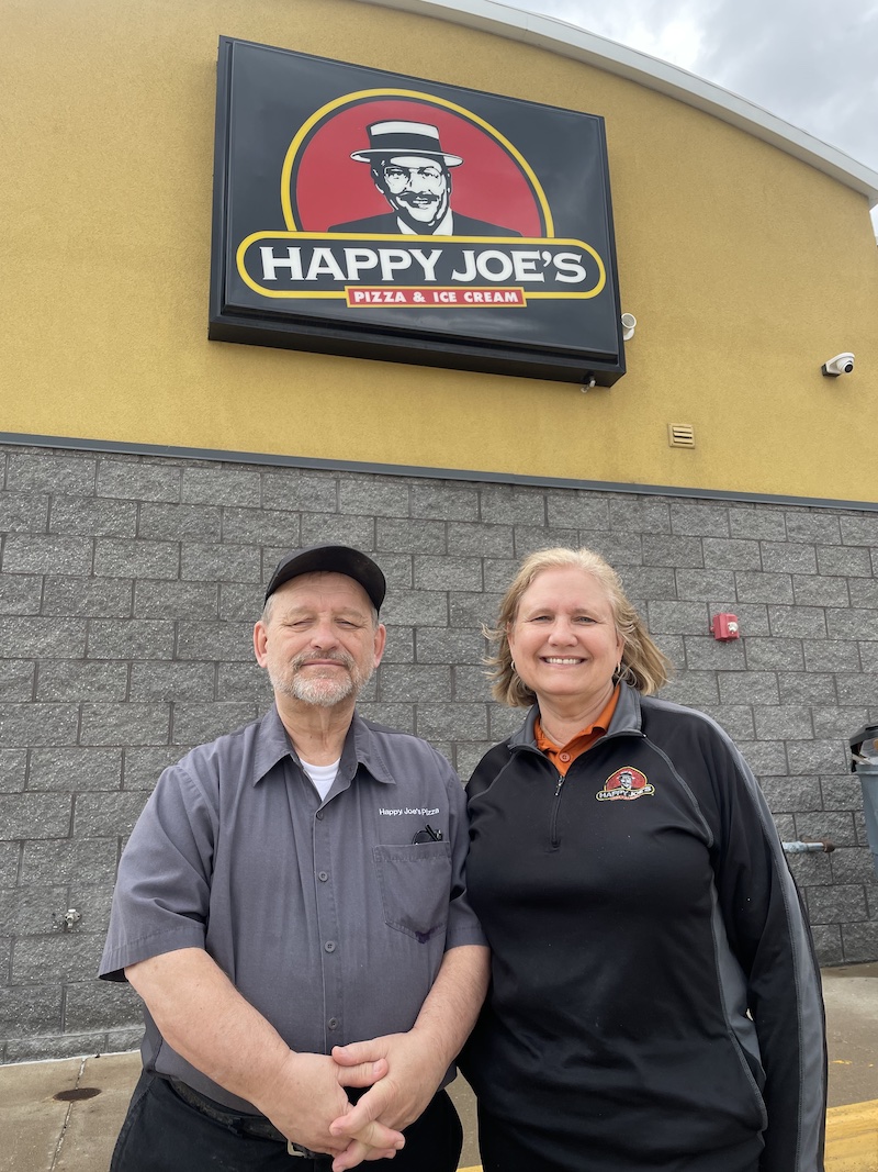 This photo shows Will and Deb Brinkley standing side by side in front of their Happy Joe's restaurant with the Happy Joe's sign looming above them.