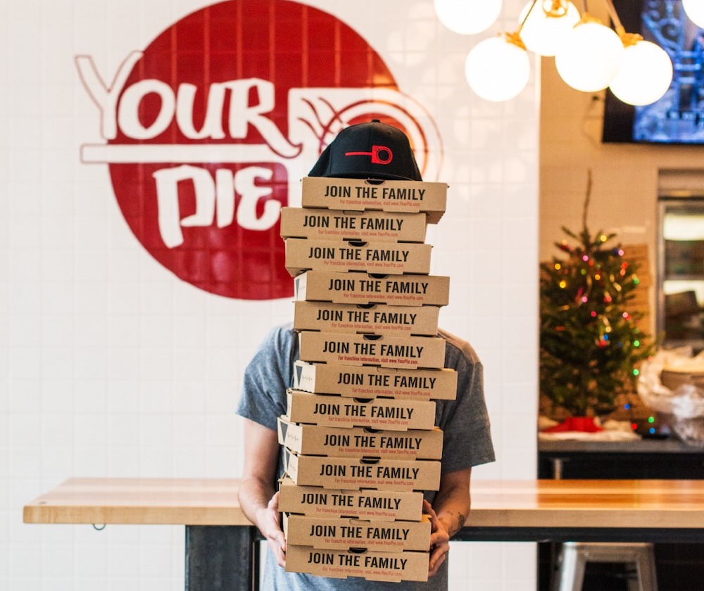 this photo shows a man standing in front of the Your Pie logo and holding a stack of delivery boxes that reaches so high, it obscures his face