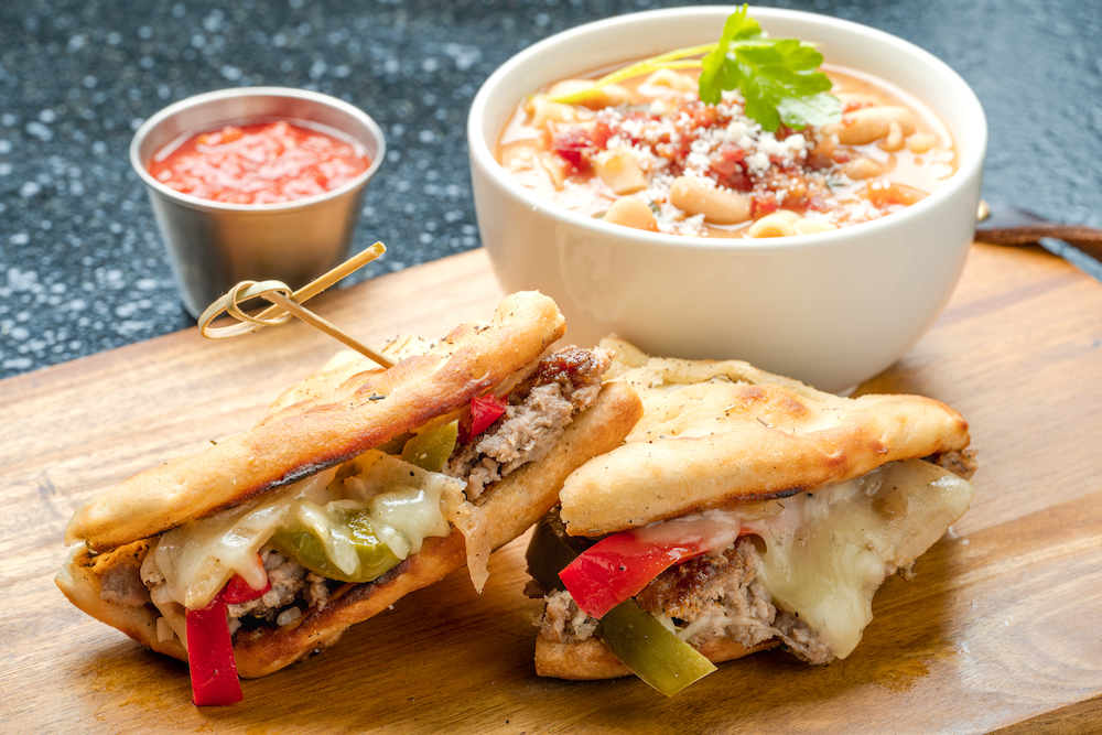 this photo shows a bowl of pasta fagoli soup paired with an Italian sausage and peppers sandwich