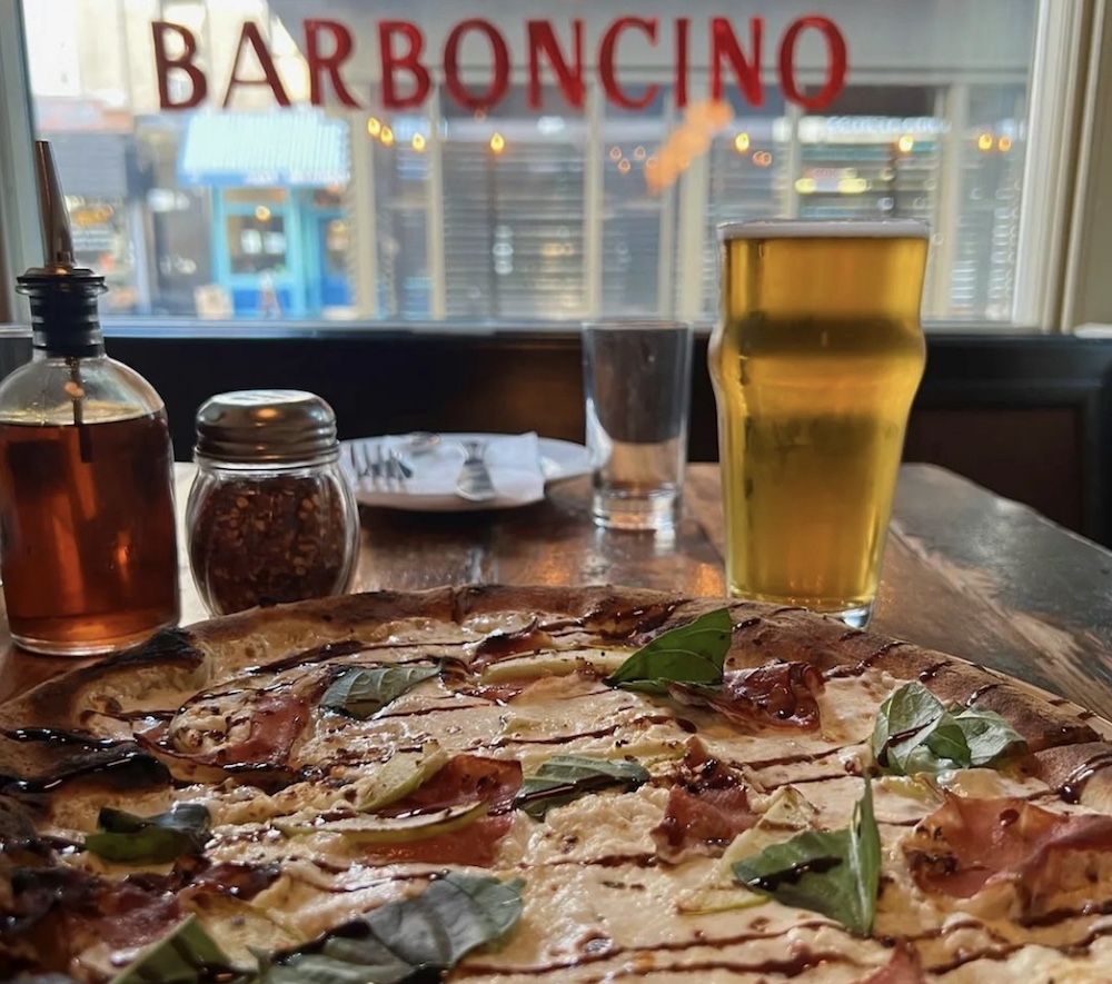 this photo shows a pizza, a glass of beer, a container of olive oil and a container of red pepper flakes in front of lettering on a window that reads Barboncino